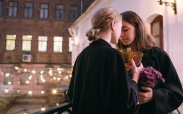 Two women facing each other in a loving moment. Both are holding a glass each and one is holding purple flowers.