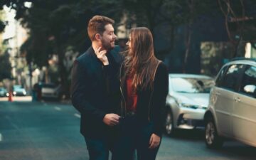 A man and women walking down a street during the day. Both are smiling looking at each other and the woman has her hand lovingly on the man's face.