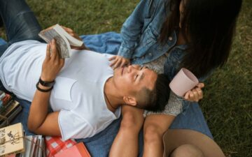 man holding a book laying on woman's lap. Both are on a picnic rug.