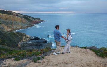 Man and woman holding hands on a cliff top with the ocean in the background